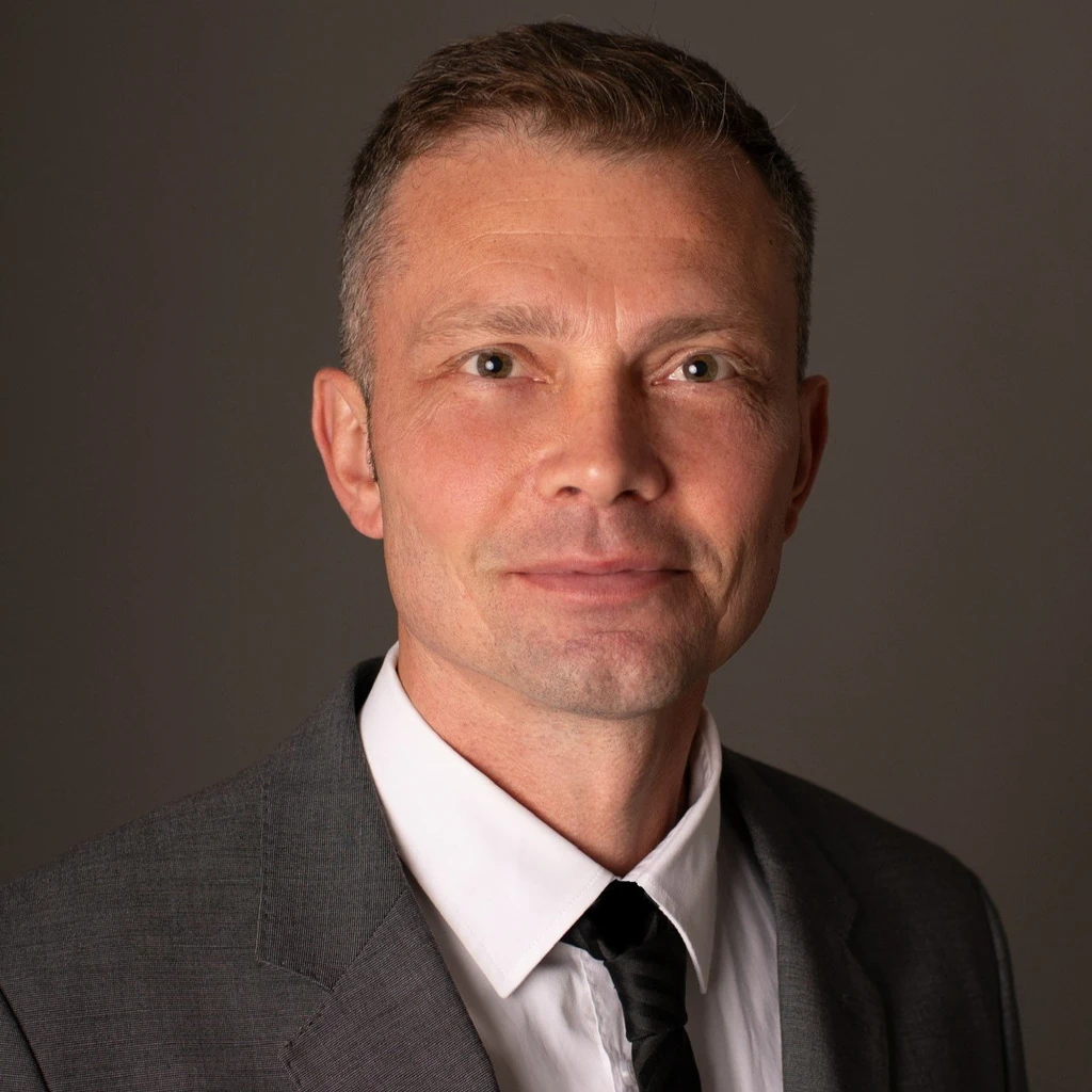 Profilbild vom Geschäftsführer Andreas Kamerer. Andreas Kamerer in einem grauen Anzug mit weißem Hemd und schwarzer Krawatte, steht vor einem neutralen dunklen Hintergrund, wirkt professionell und seriös
